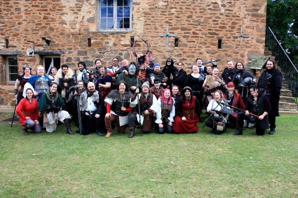 A group of people in medieval-inspired costume kneel and stand in a posed photograph smiling at the camera. A stone wall stands behind them.